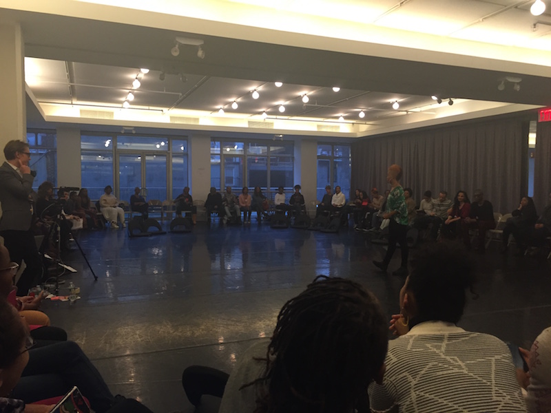 A group of woman sit in folding chairs around the perimeter of a studio as Camille A. Brown talks in the center